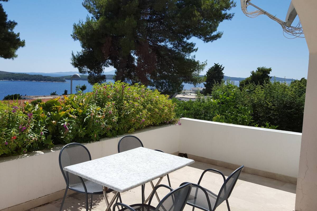A terrace with a table and chairs, overlooking a coastal view with trees and a clear sky.