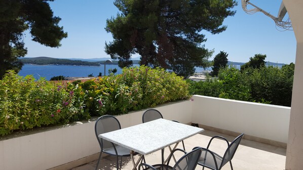 A terrace with a table and chairs, overlooking a coastal view with trees and a clear sky.