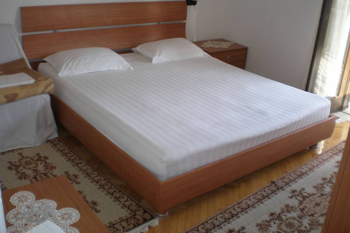 A neatly made bed with white linens in a room with wooden flooring and a patterned rug.