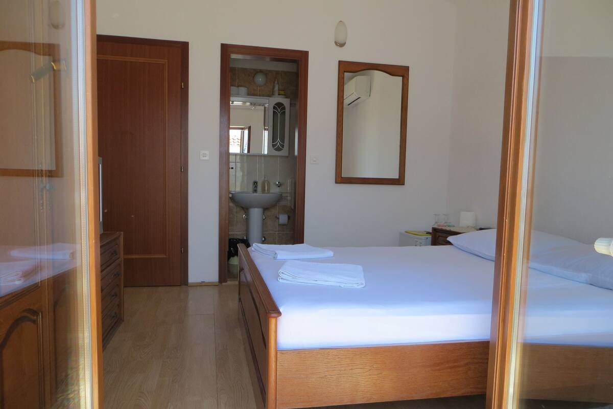 A bedroom with a wooden bed, a bedside table, a mirror, and a bathroom visible through an open door.