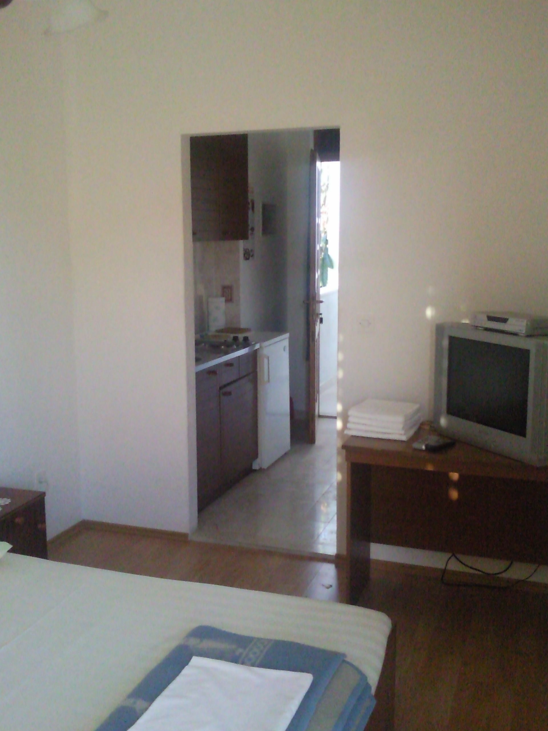 A bedroom with a bed, a TV on a wooden table, and a kitchen in the background.