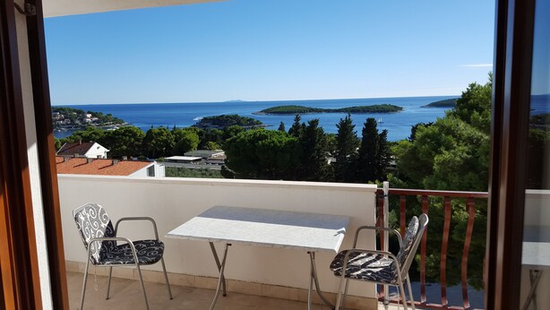 A balcony with a table and two chairs, overlooking a coastal view with houses and trees. - Apartments and rooms Rogosic (Hvar)