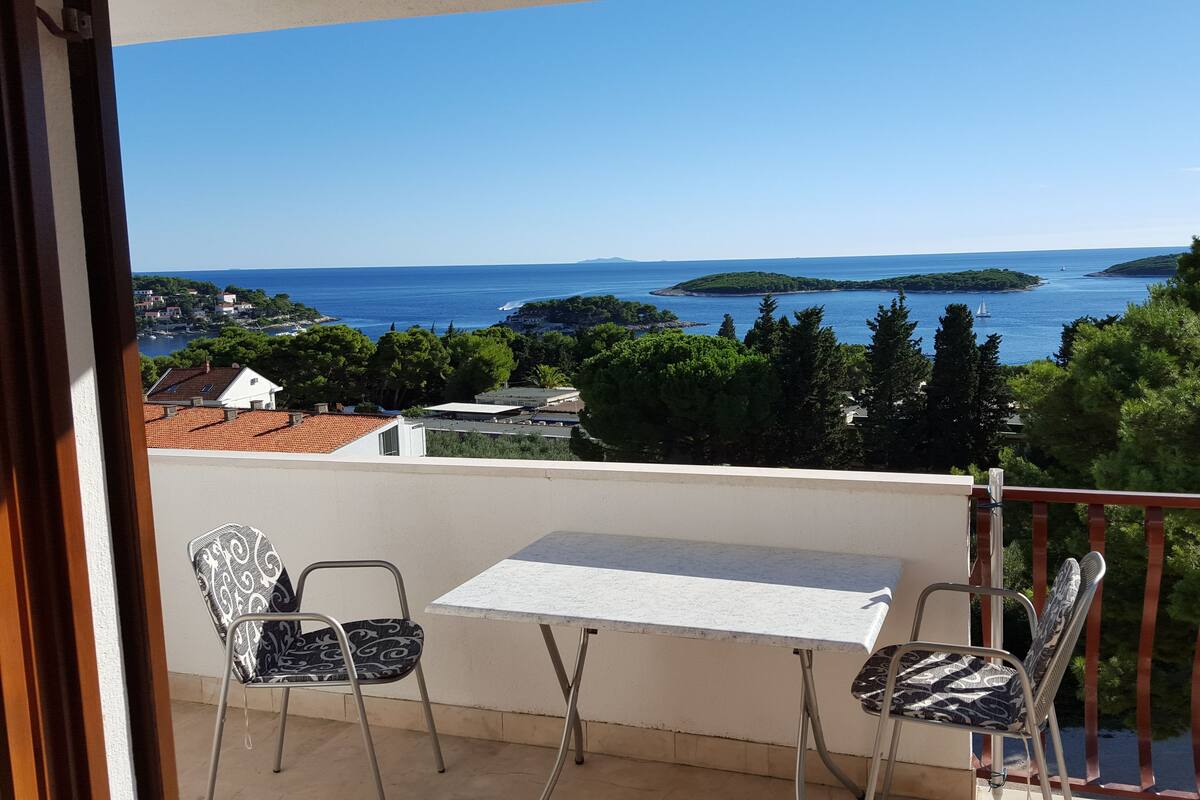 A balcony with a table and two chairs, overlooking a coastal view with houses and trees.