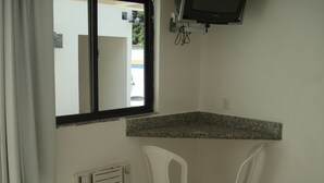 A small kitchenette with a granite countertop, two white plastic chairs, a window with a black frame, and a wall-mounted television. - Pousada Praia da Ribeira Clube (Angra dos Reis)