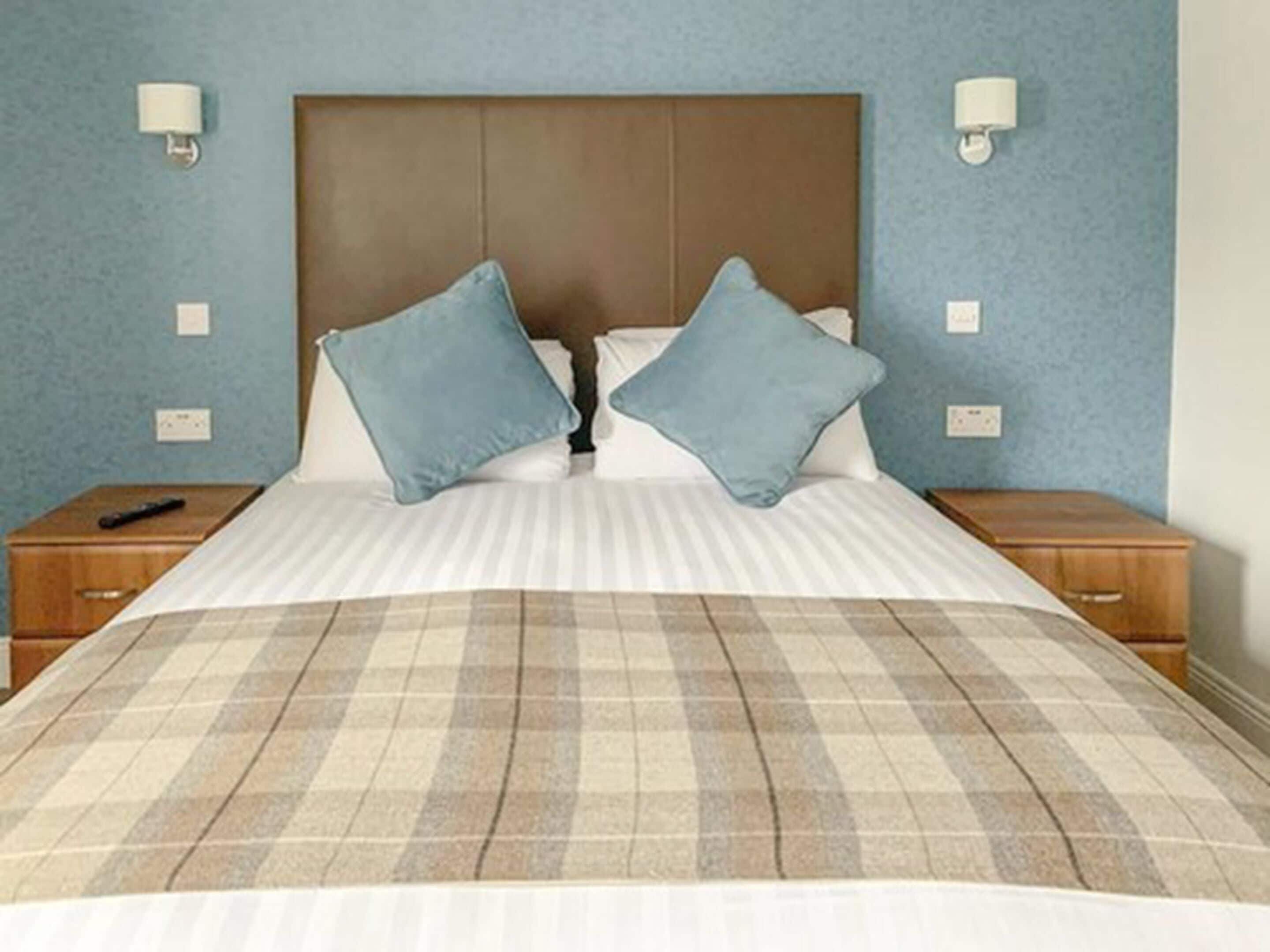 Una cama con colcha escocesa, dos almohadas y un cabecero de madera. Hay dos mesitas de noche, una con lámpara y control remoto.