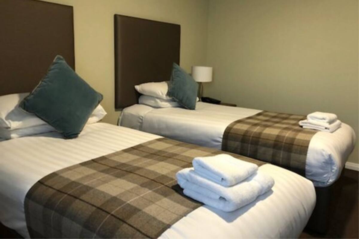 A hotel room with two beds, each with a headboard, a lamp, and a plaid blanket.