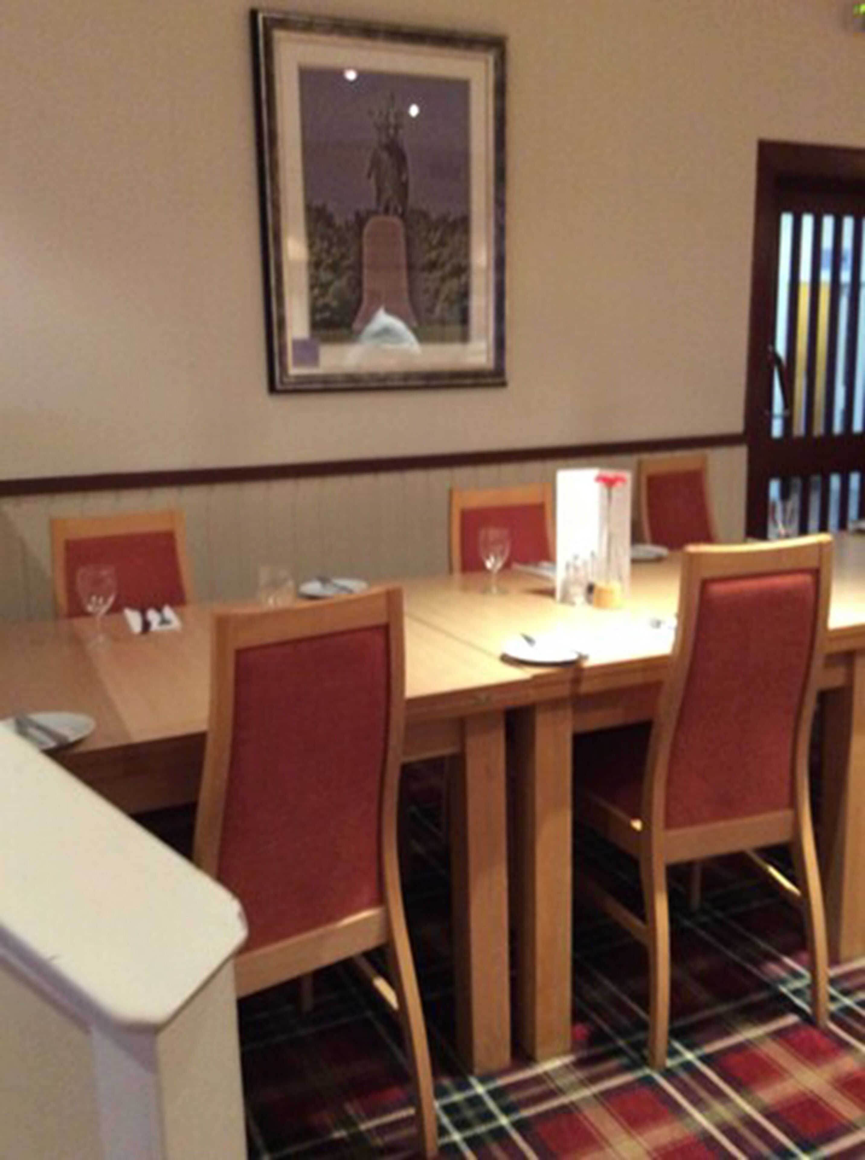 A dining area with a wooden table and red upholstered chairs, a framed picture on the wall, and a plaid carpet.