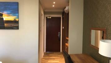 A hotel hallway with a wooden floor, a bed with a white bedspread, a wooden desk with a lamp, and a painting on the wall.