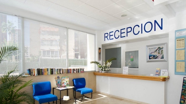 A reception area with a sign that reads "RECEPCION," a reception desk, two blue chairs, a small table with a book, and a plant. - Benimar (Benidorm)