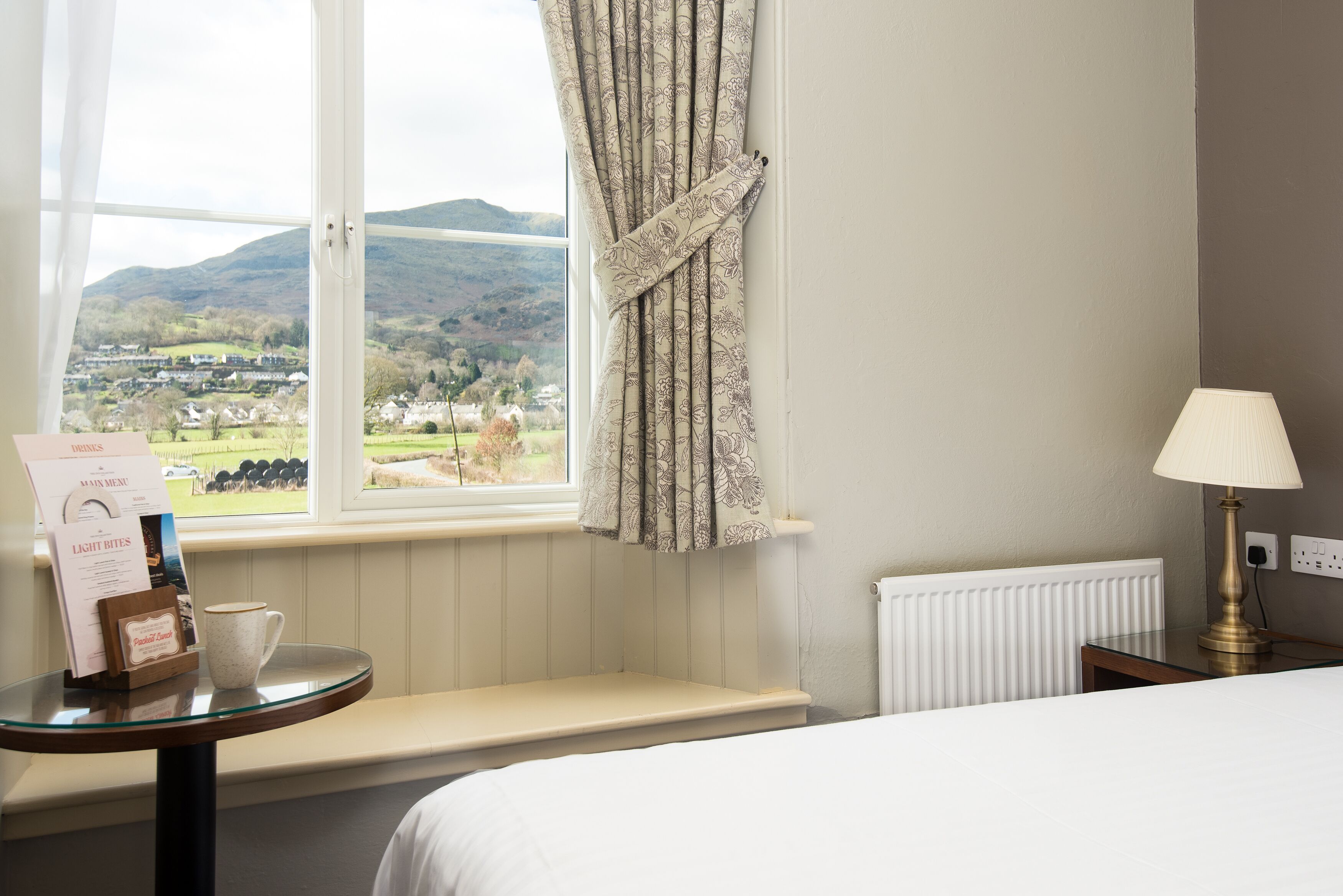 A hotel room with a large window offering a scenic view of a valley, a bedside table with a menu and a mug, and a lamp on the nightstand.