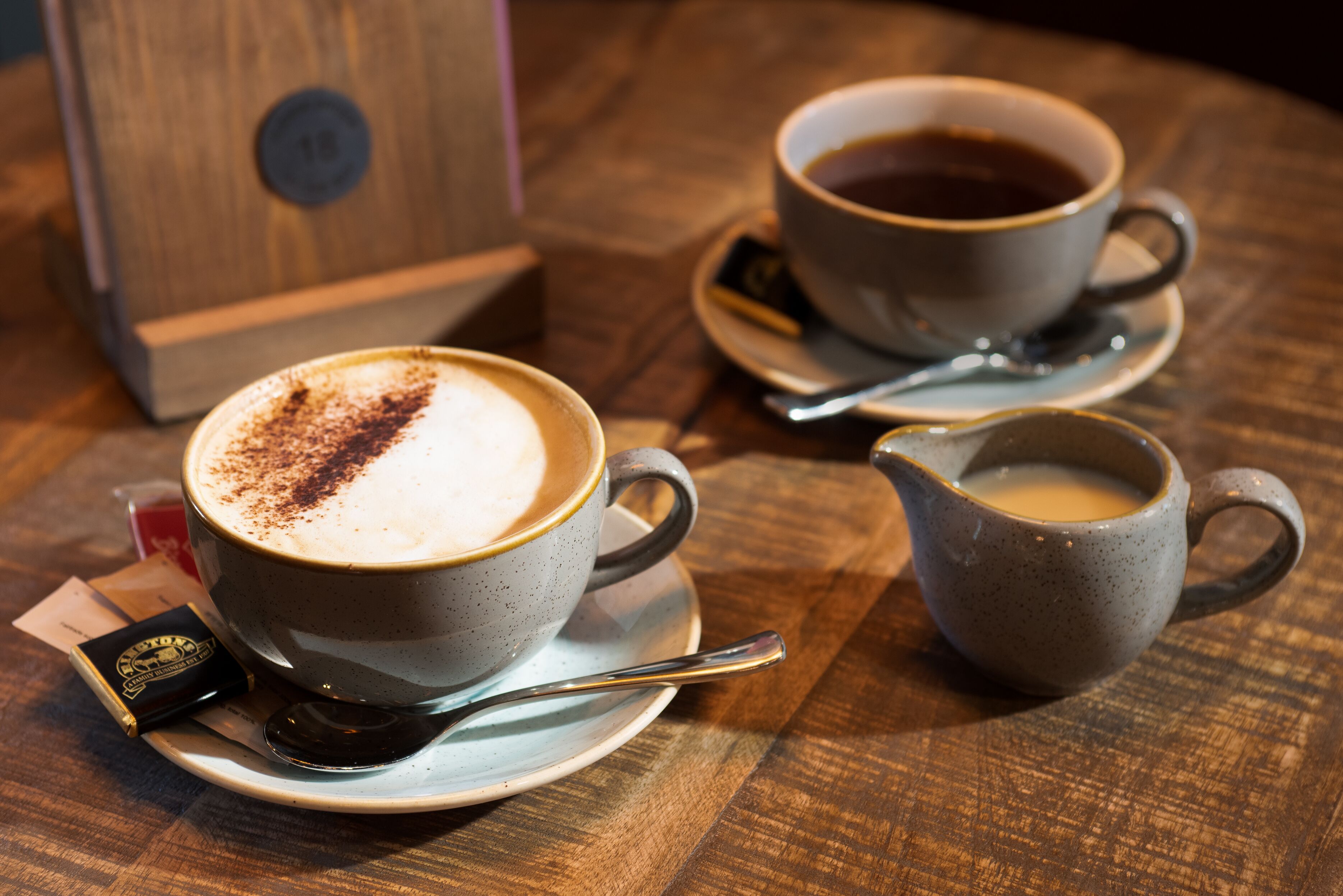 Two cups of coffee on saucers with spoons, a small pitcher, and a wooden box on a wooden table.