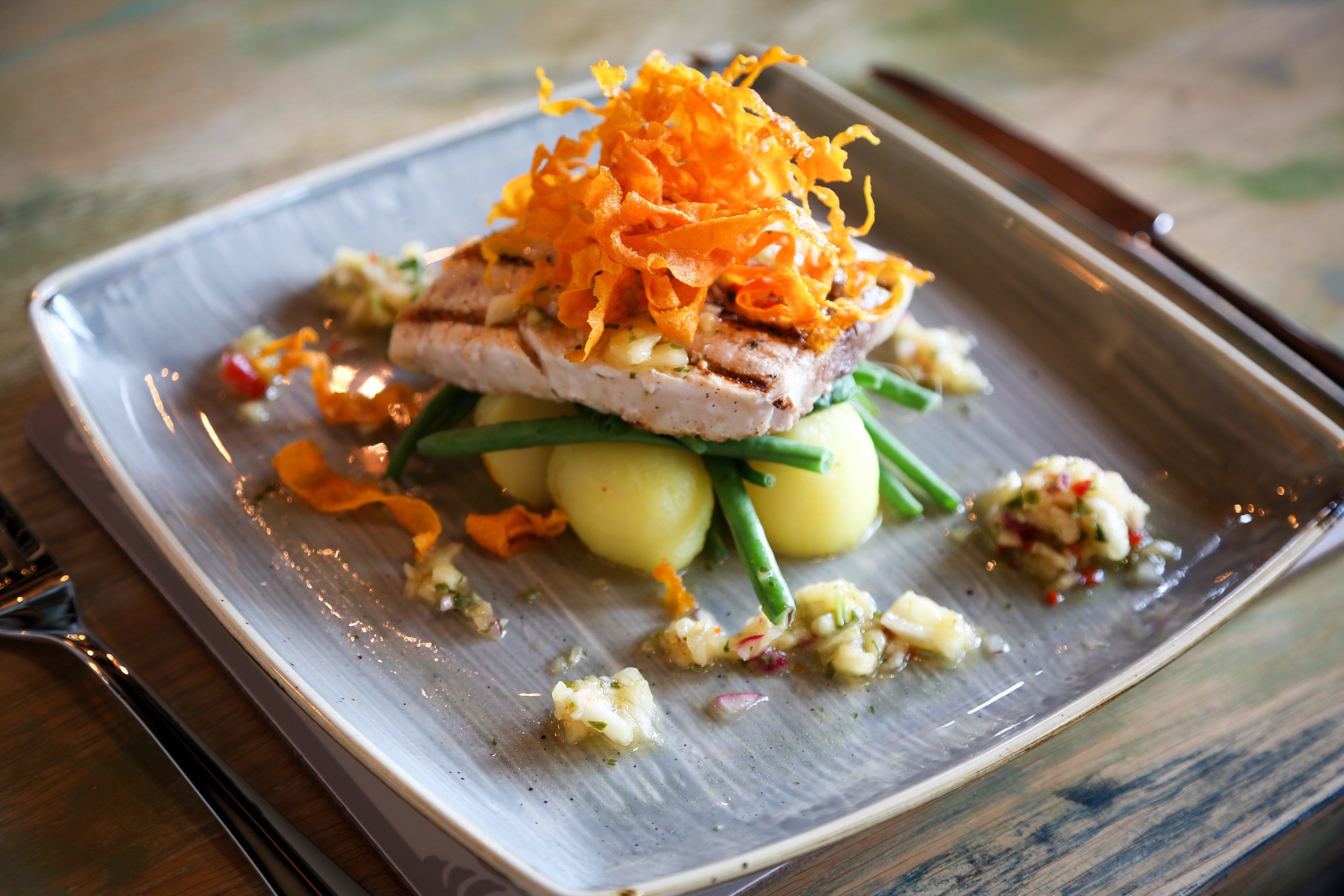 A square plate with a grilled fish topped with shredded orange vegetable, green beans, and a side of mashed potatoes.