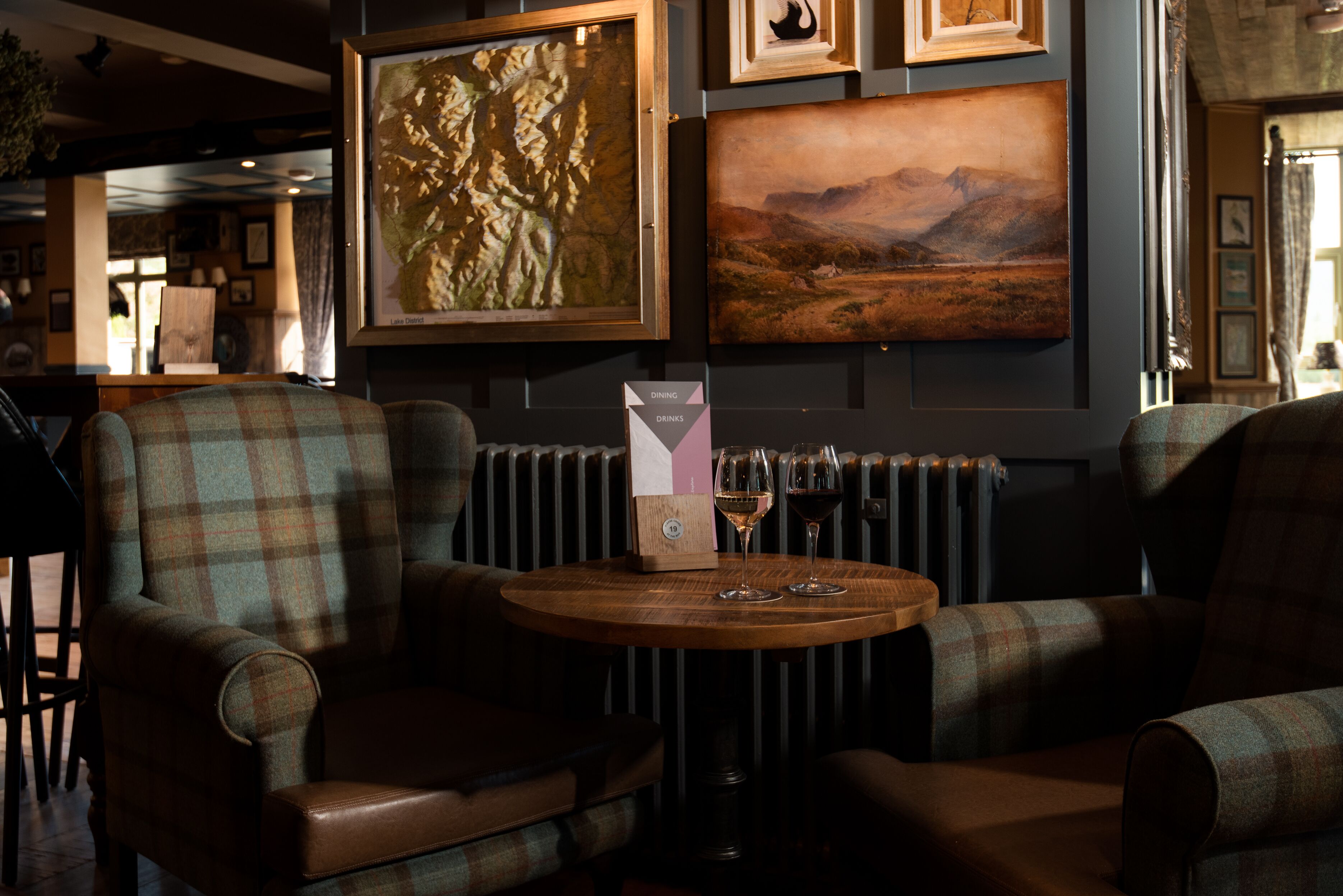 A cozy seating area with plaid upholstered chairs and a wooden table, featuring framed artwork on the wall and a radiator below.