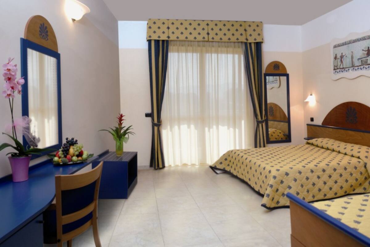 Una habitación de hotel con una cama, un escritorio con una silla, una mesita con frutero y una planta en maceta, una ventana con cortinas y un espejo montado en la pared.