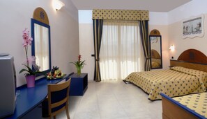 A hotel room with a bed, a desk with a chair, a small table with a fruit bowl and a potted plant, a window with curtains, and a wall-mounted mirror. - Akiris (Nova Siri)