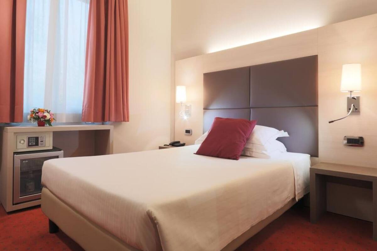 A hotel room with a large bed, a bedside table with a lamp, a small desk with a computer, and a window with curtains.