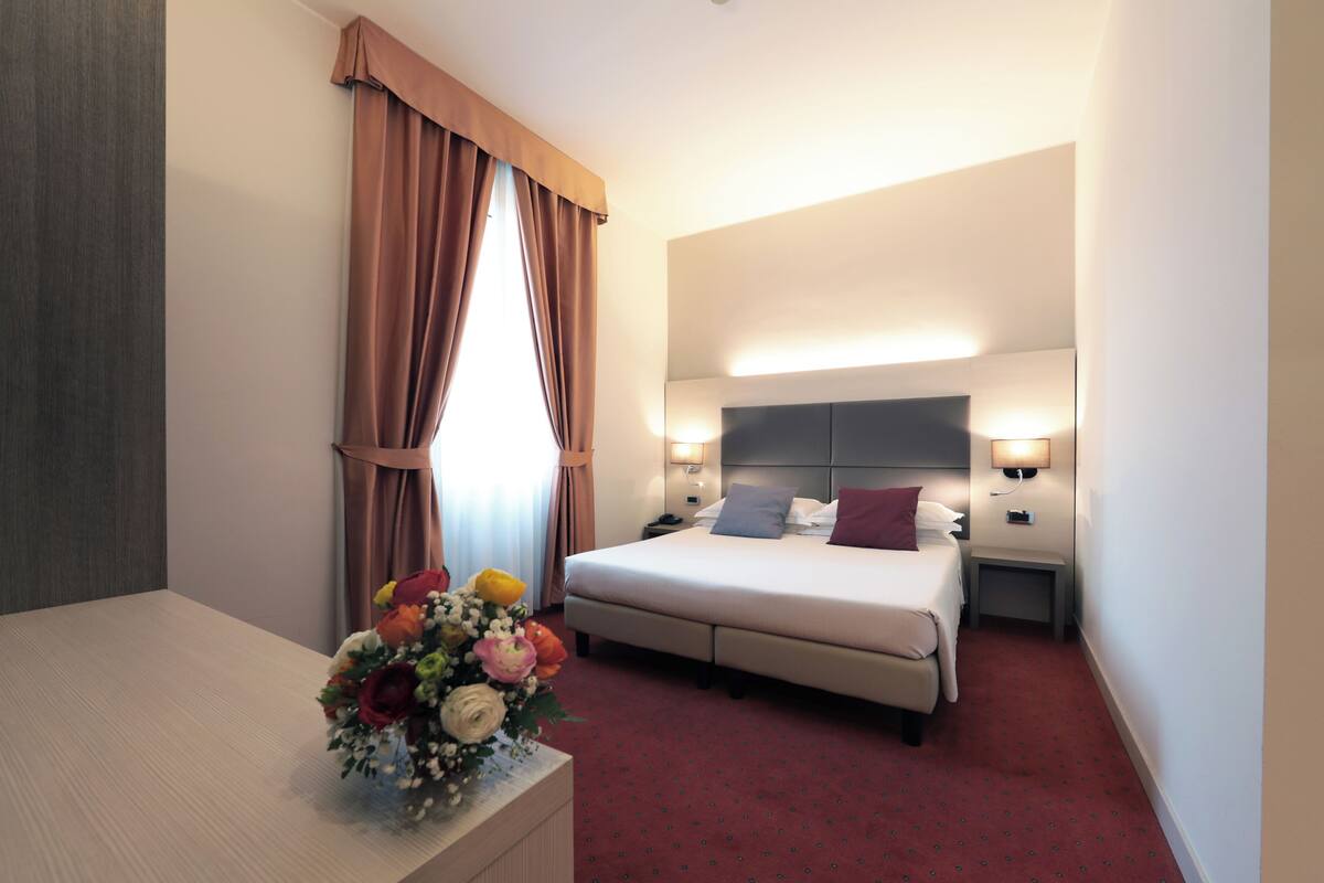 A hotel room with a bed, bedside tables, a desk, and a flower arrangement.