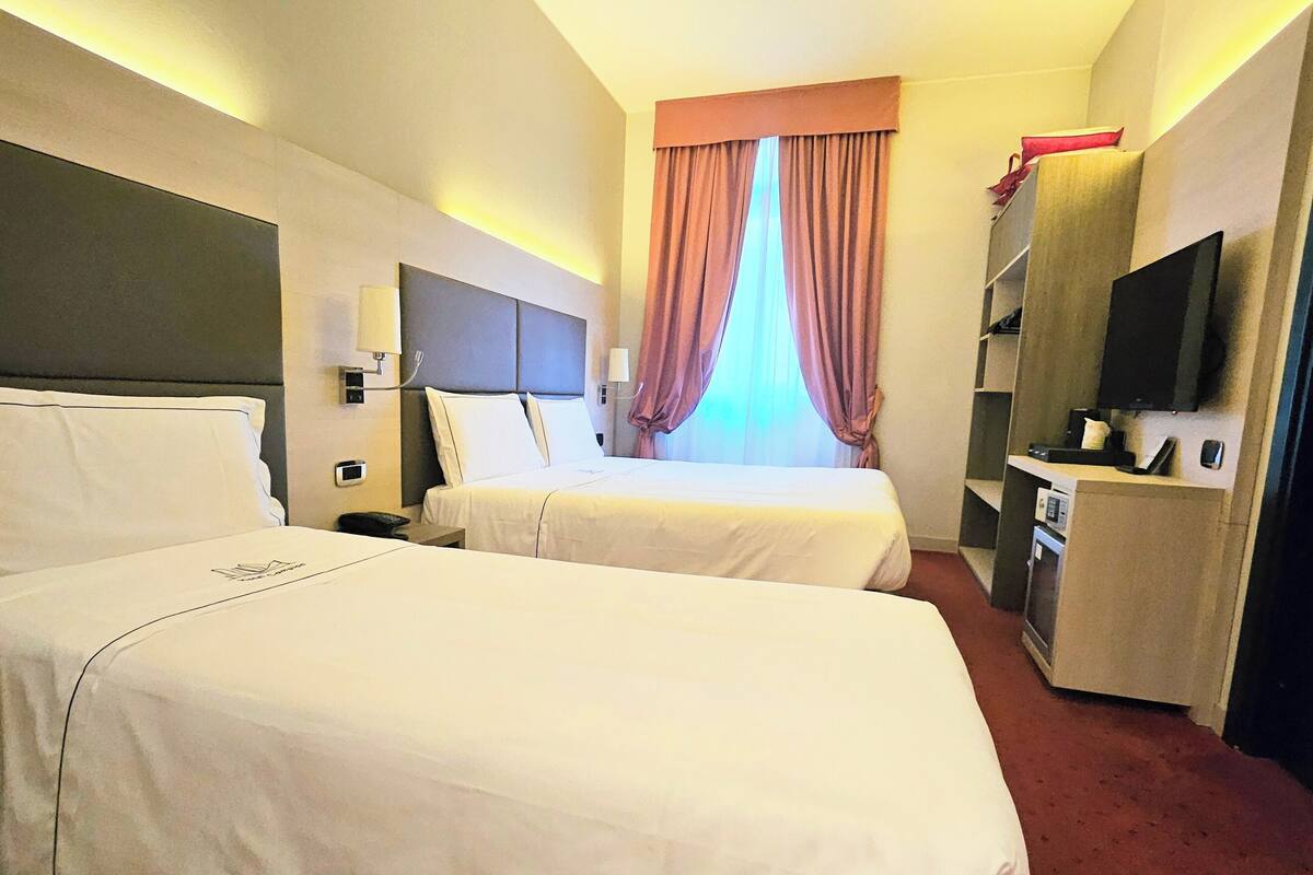 A hotel room with two beds, a television, a desk, and a window with curtains.