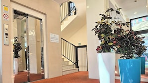 Un vestíbulo de hotel con un ascensor de vidrio, una escalera y grandes plantas en macetas.