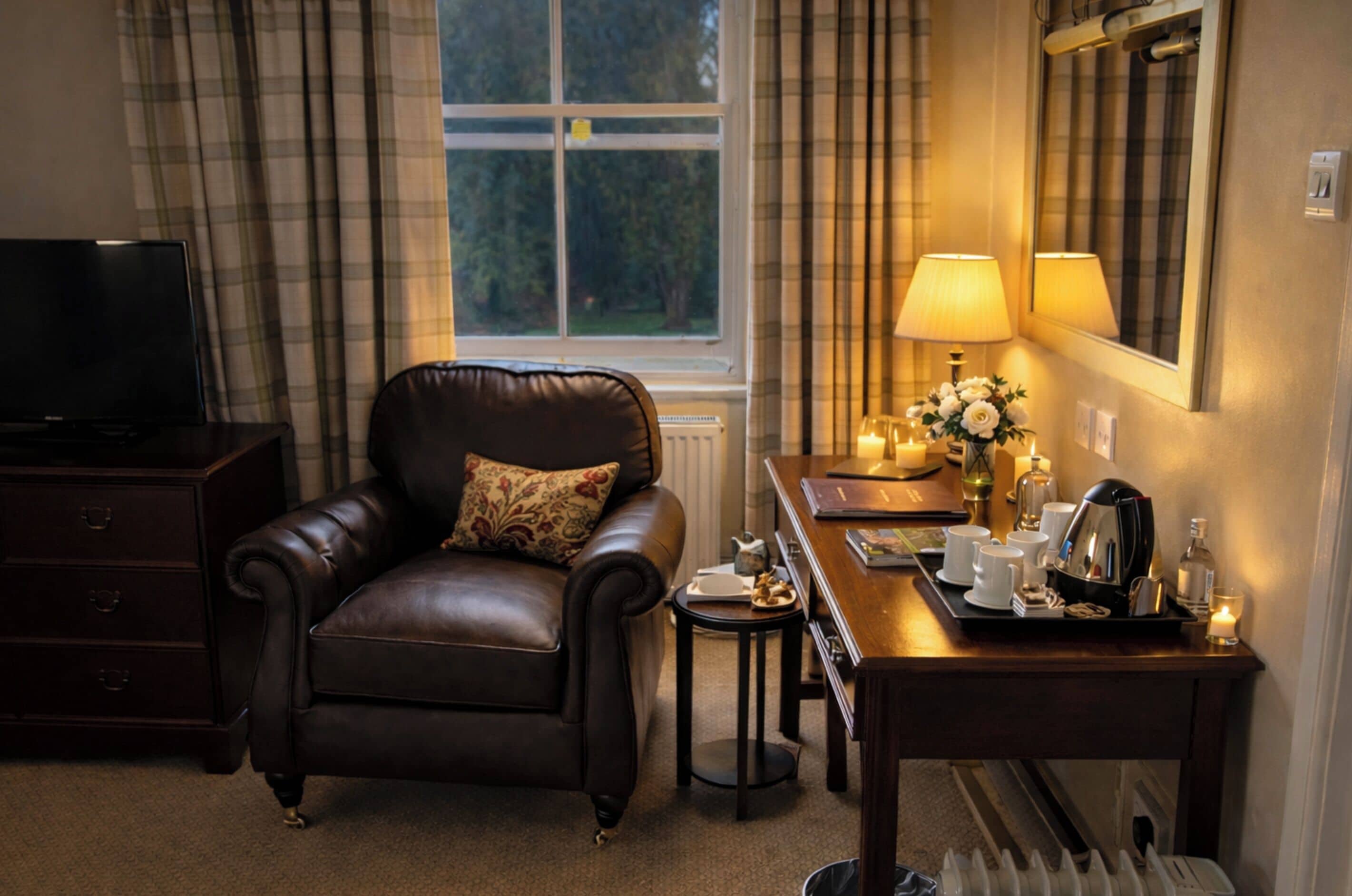 A cozy hotel room with a leather armchair, a wooden desk with a lamp, a vase of flowers, and a view of trees through a window.