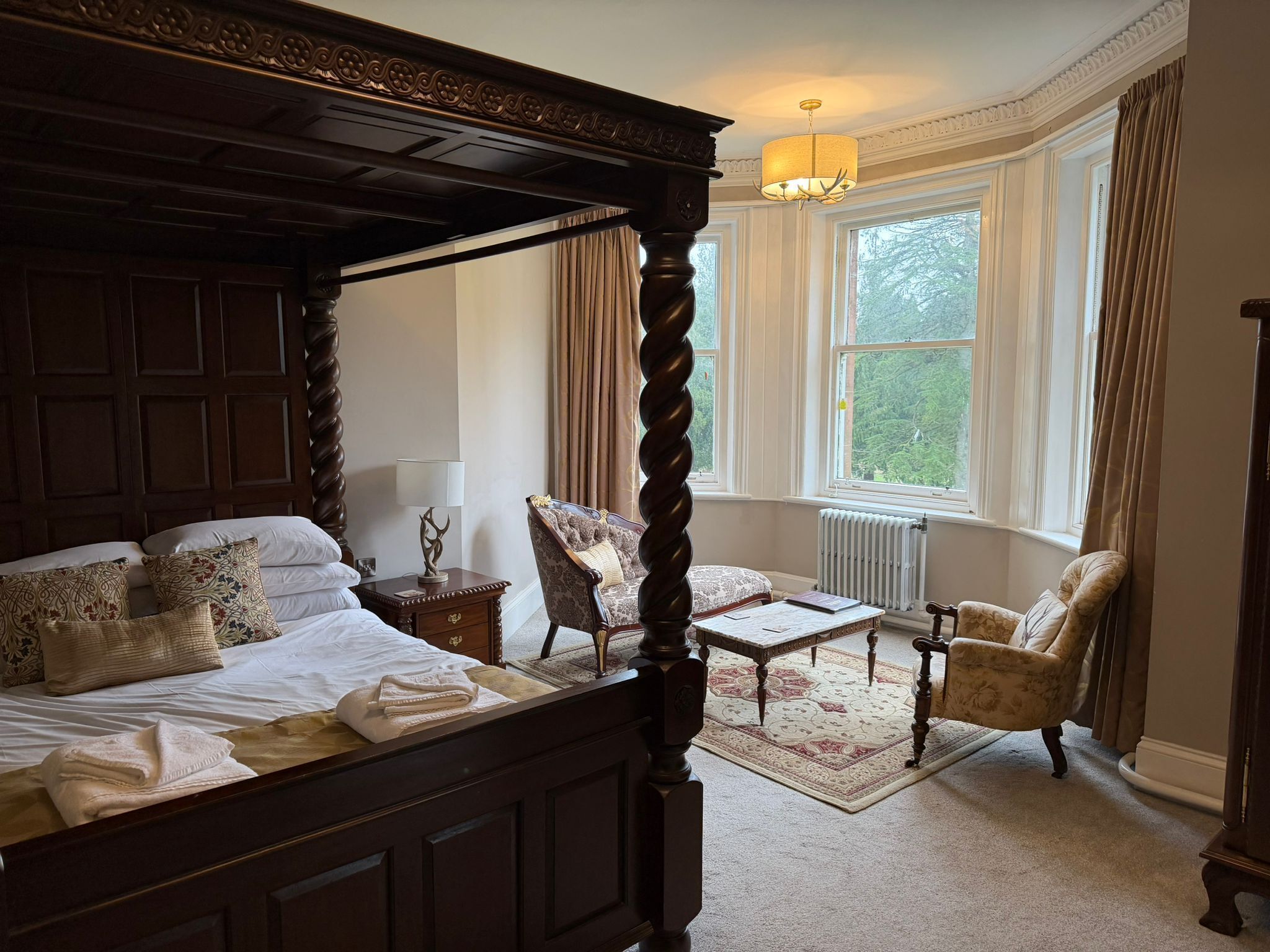 Un lit à baldaquin avec une tête de lit en bois, une table de chevet avec une lampe, une chaise et une petite table dans une pièce dotée de grandes fenêtres et d’un radiateur.