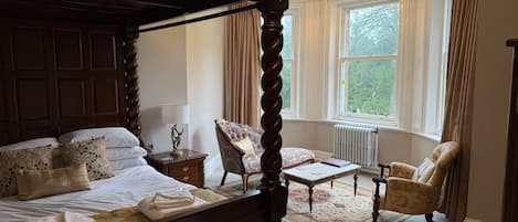 Ein Bett mit Baldachin, ein Holzkopfstück, ein Nachttisch mit Lampe, ein Sessel und ein kleiner Tisch in einem Zimmer mit großen Fenstern und einer Heizung.