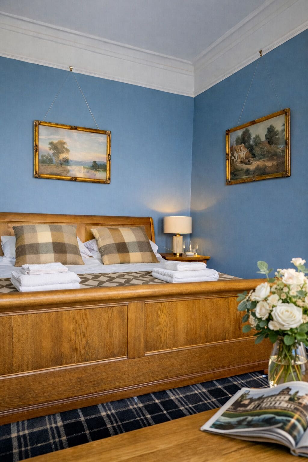 A neatly made bed with checkered pillows and folded towels, a bedside lamp, and two framed paintings on the wall.