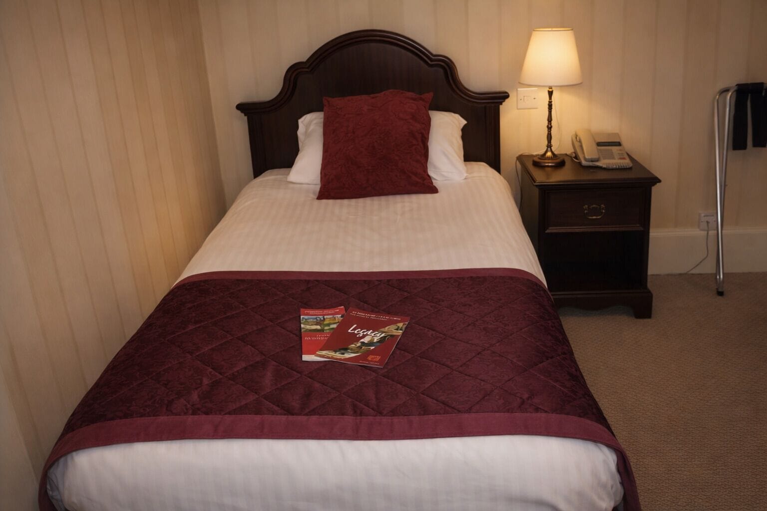 A neatly made bed with a red quilt and pillows, a bedside lamp, and a nightstand with a phone.
