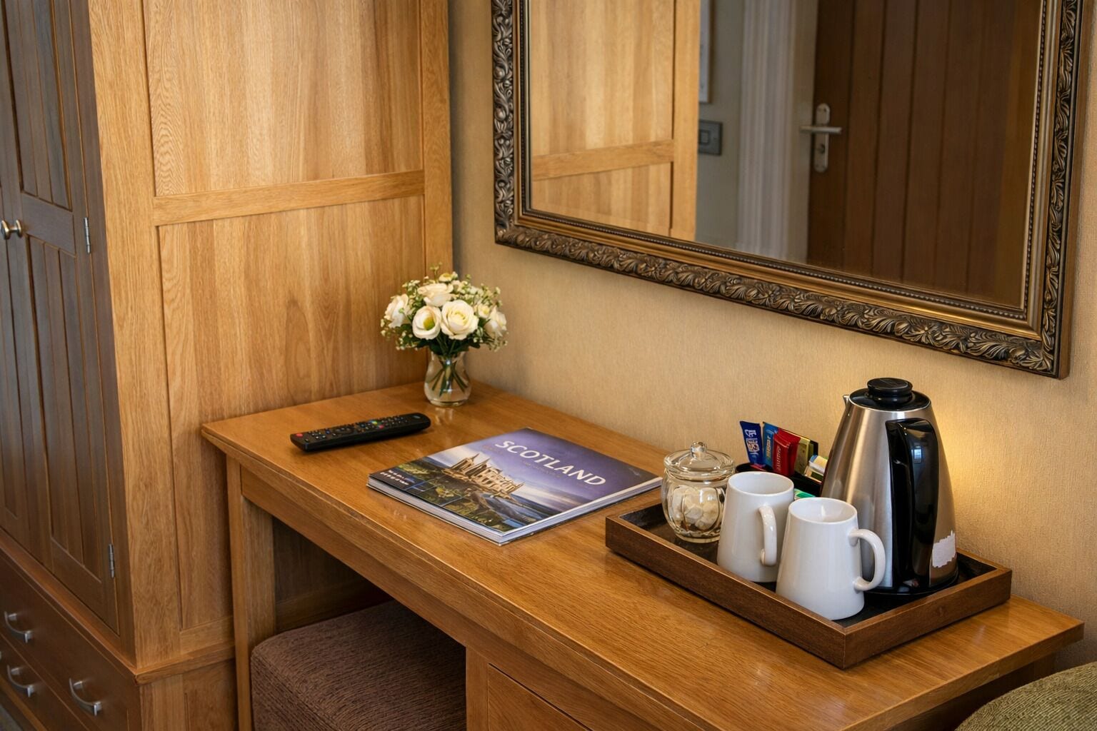 Un escritorio de madera con un control remoto, una revista de viajes, una cafetera y una bandeja con tazas y un vaso.