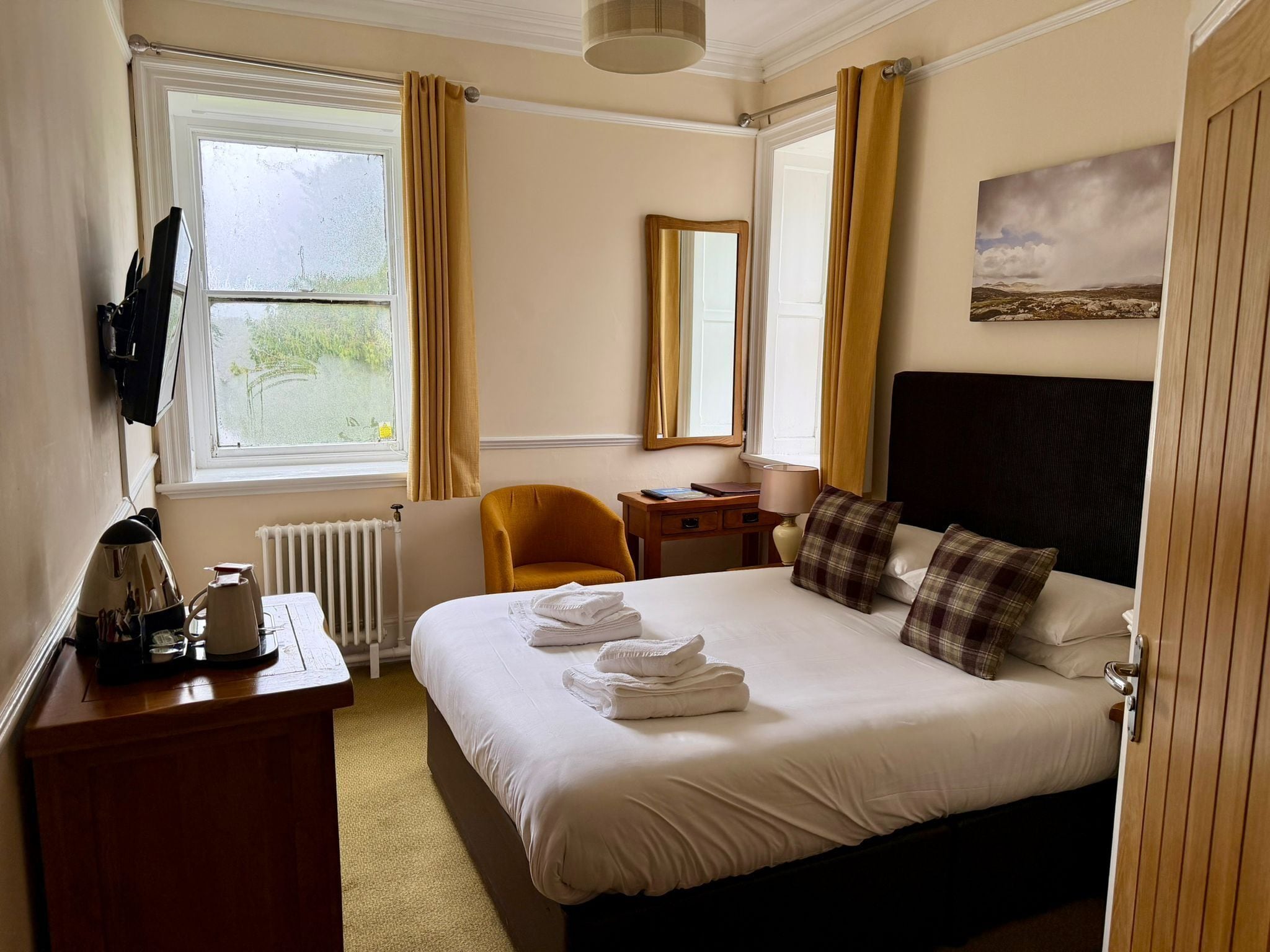 A hotel room with a bed, a desk, a chair, a radiator, a television, and a painting on the wall.