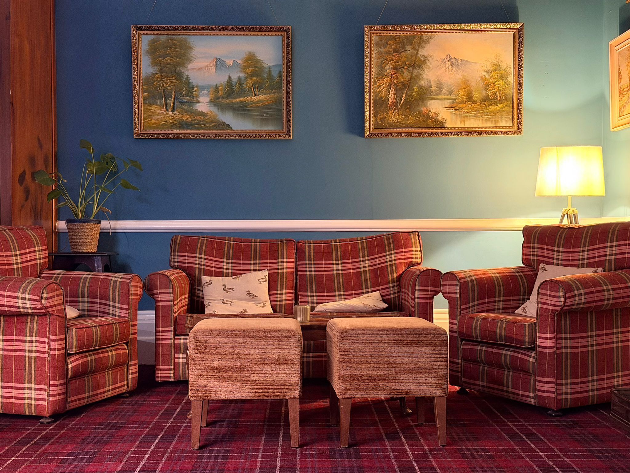 A room with a blue wall, two framed landscape paintings, a plaid sofa set, a small table with a lamp, and a potted plant.