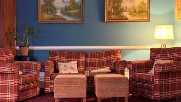 A room with a blue wall, two framed landscape paintings, a plaid sofa set, a small table with a lamp, and a potted plant.
