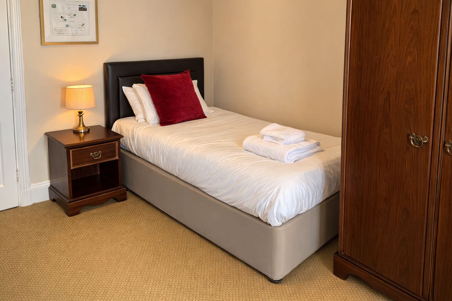 Una cama bien tendida con sábanas blancas y un cojín rojo, una lámpara de noche, una mesita de noche de madera con cajón, y un armario de madera.