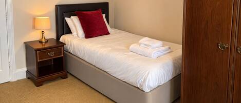 Una cama bien tendida con sábanas blancas y un cojín rojo, una lámpara de noche, una mesita de noche de madera con cajón, y un armario de madera.