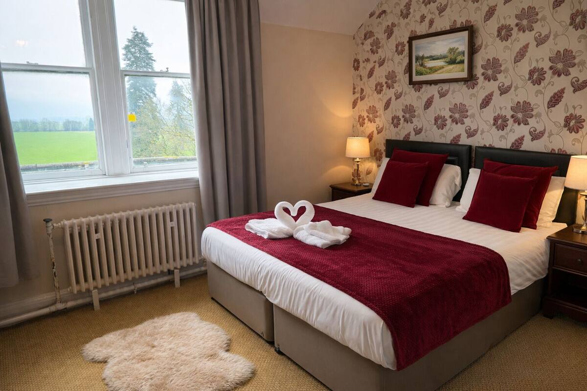 A hotel room with a large bed, a radiator, a window with curtains, a fluffy rug, and a floral wallpaper.