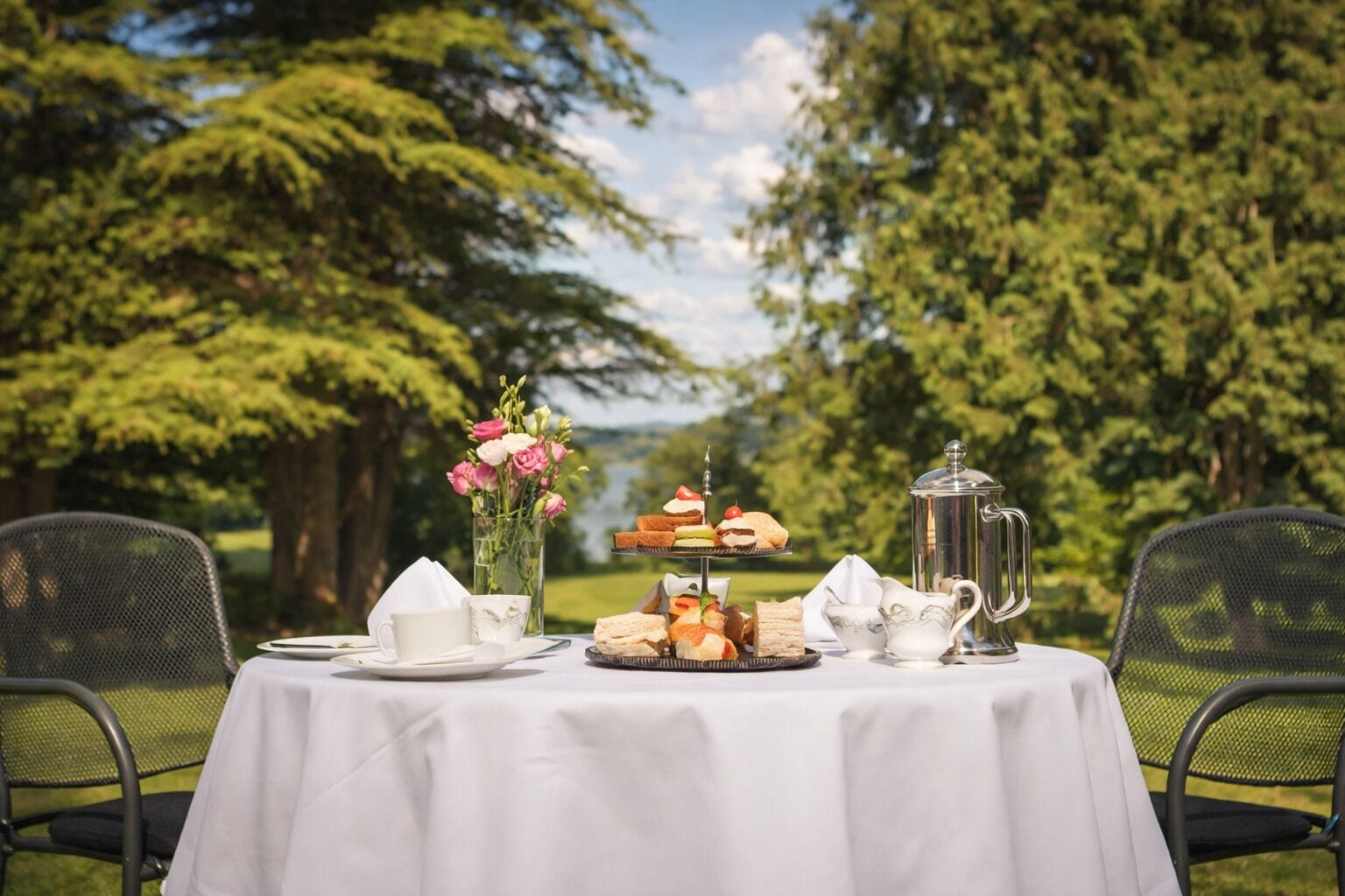 Mesa al aire libre con mantel blanco, una jofaina, tazas y un pastelero escalonado.