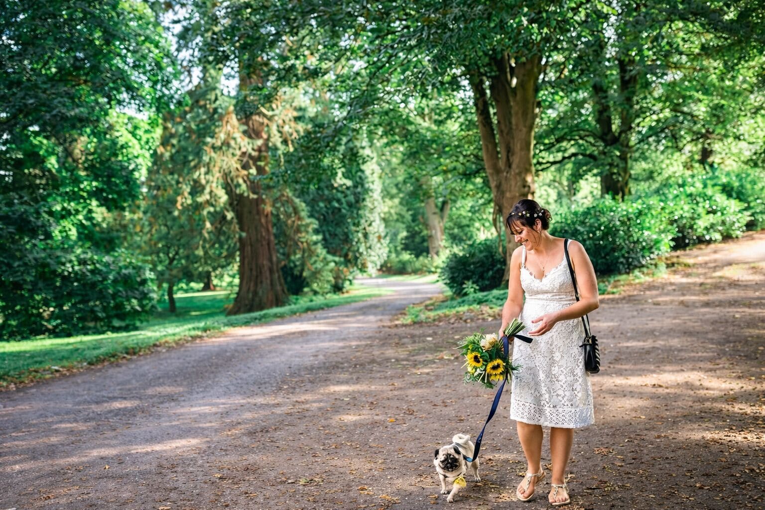 A woman in a white dress walking a small dog on a leash, holding a bouquet of sunflowers, in a park with trees and a dirt path.