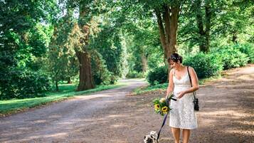 A woman in a white dress walking a small dog on a leash, holding a bouquet of sunflowers, in a park with trees and a dirt path.