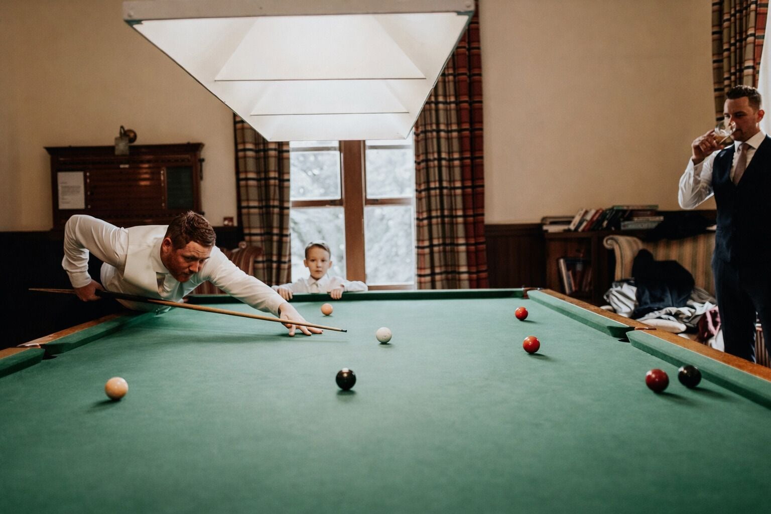 A pool table in a room with a large window, a lamp, and a bookshelf.