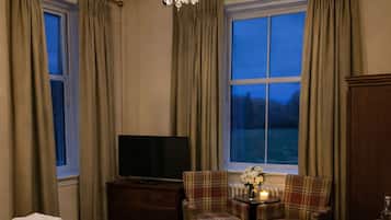 A hotel room with a large bed, a TV, a chandelier, a table with chairs, and a view of the outdoors.