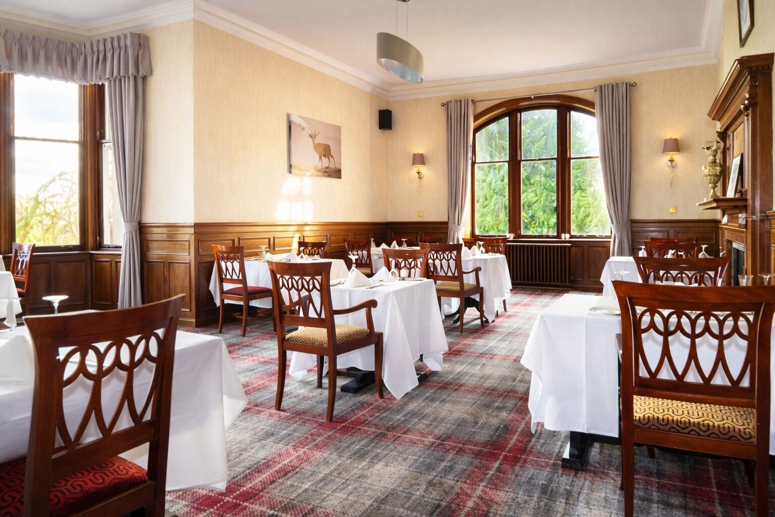 A dining area with wooden chairs and tables, a plaid carpet, and large windows with curtains.