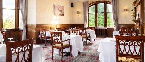 A dining area with wooden chairs and tables, a plaid carpet, and large windows with curtains.