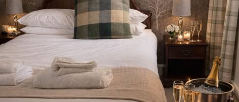 A neatly made bed with white linens, a green and white checkered throw pillow, and a bottle of champagne in a bucket. There are two lit candles on the bedside table and a glass of champagne on the floor.