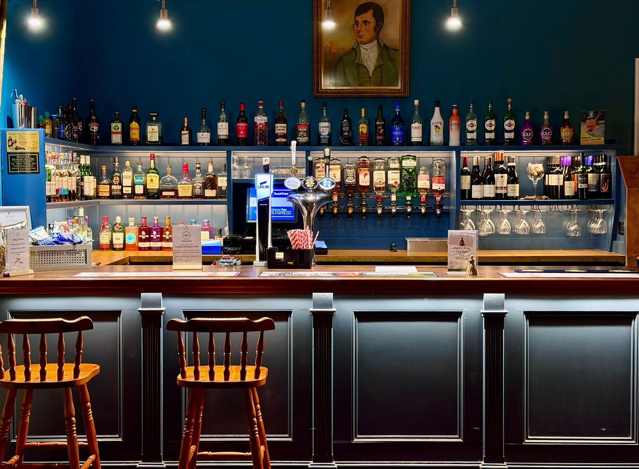 Un bar avec une variété de bouteilles d’alcool, une caisse enregistreuse et deux tabourets de bar en bois.