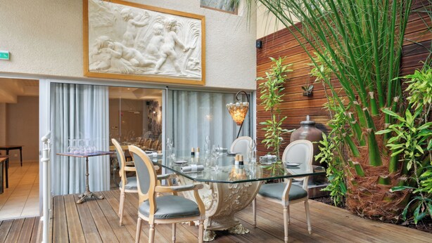 Una mesa de comedor con cubierta de vidrio, sillas, una pieza decorativa en la pared y una planta de bambú.