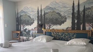 Habitación de hotel con una cama grande, cabecera con motivos florales, un mural en la pared que representa un paisaje y una silla junto a la ventana.