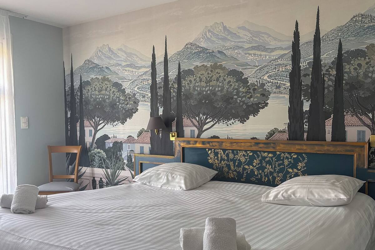 Een hotelkamer met een groot bed, een hoofdeinde met bloemenmotieven, een muurschildering van een landschap en een stoel bij het raam.