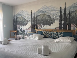 Habitación de hotel con una cama grande, cabecera con motivos florales, un mural en la pared que representa un paisaje y una silla junto a la ventana.