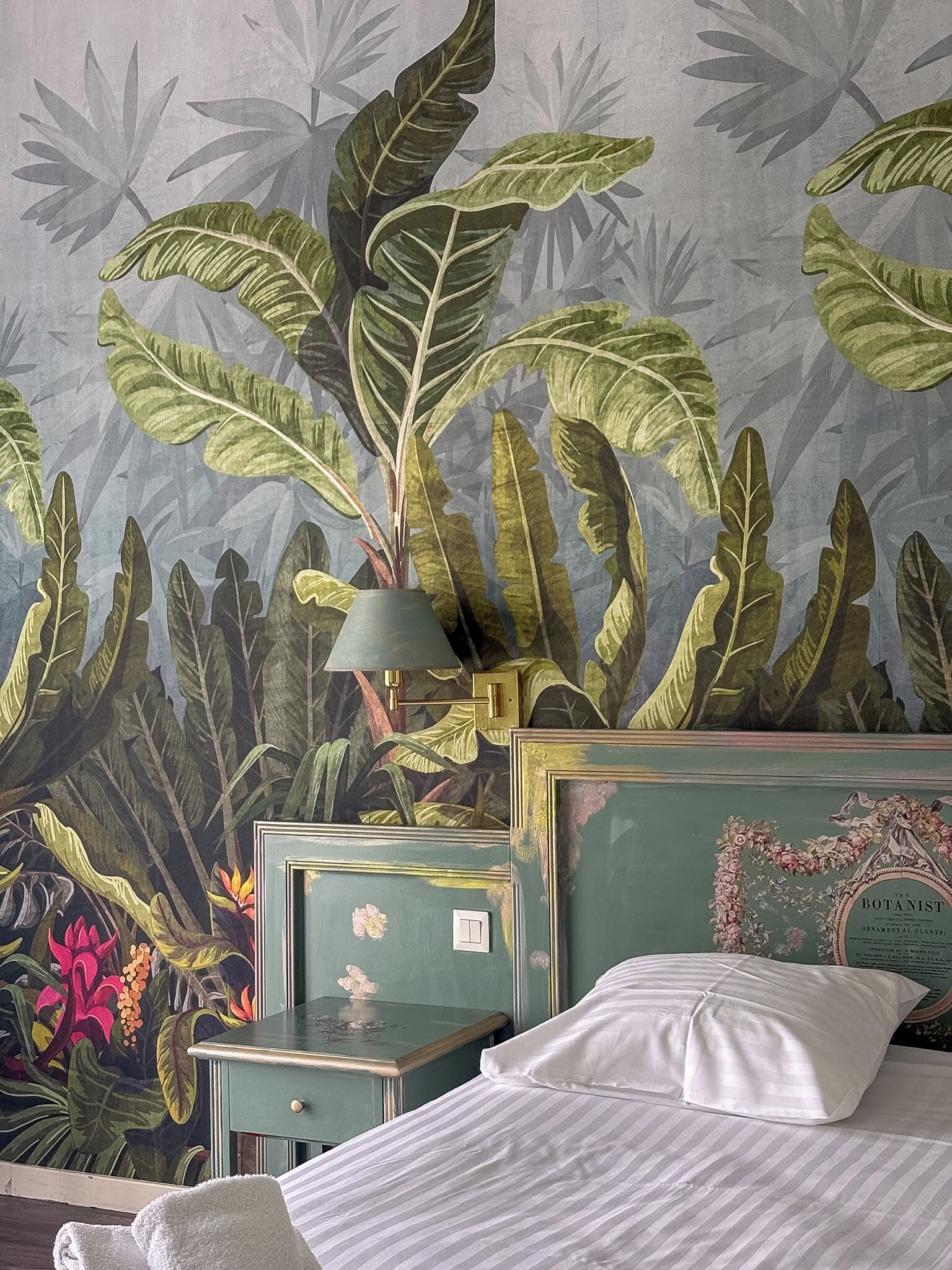 Une chambre comprenant un lit, une table de chevet, une lampe et un mur orné d’une fresque représentant des motifs végétaux.