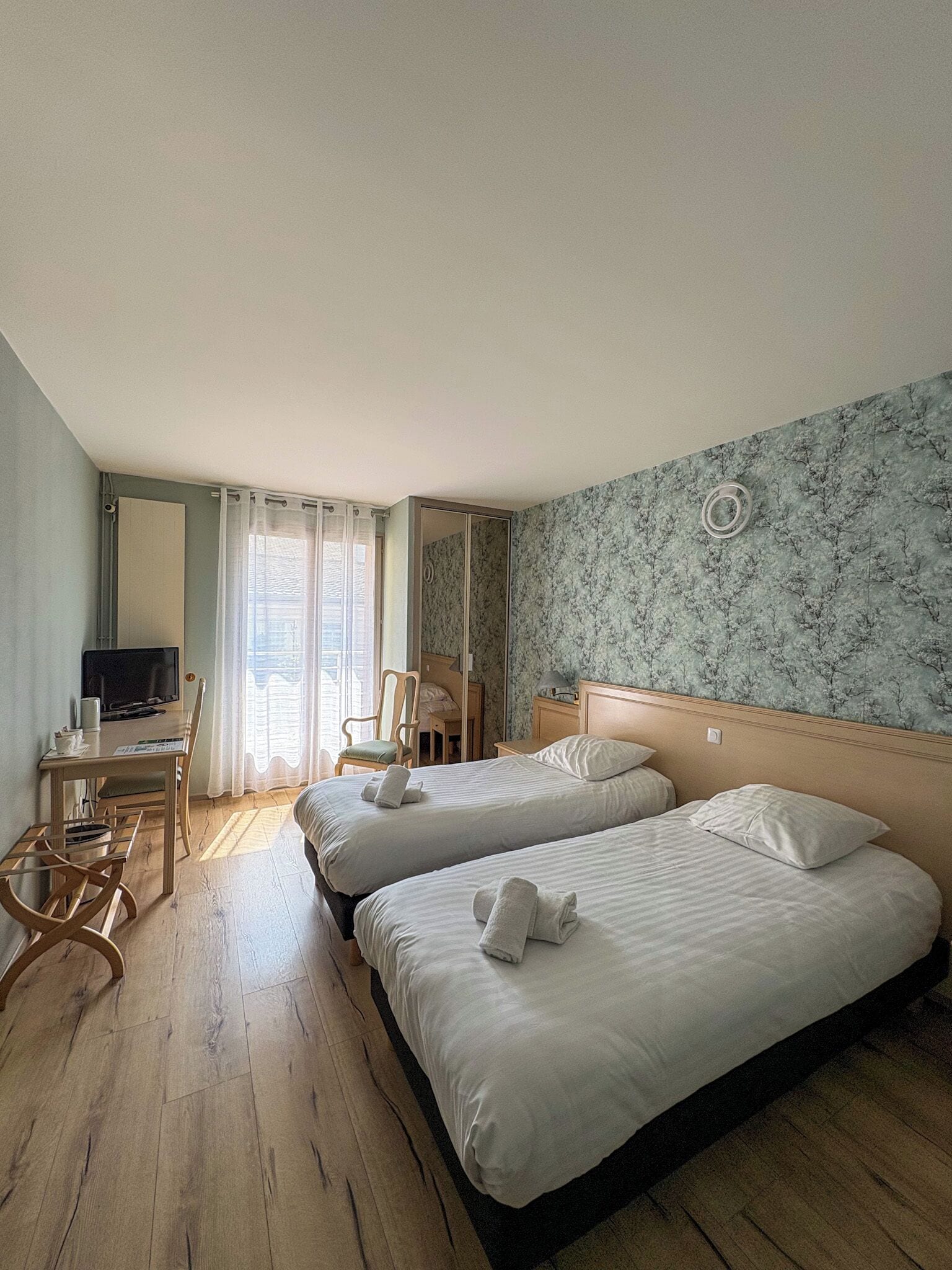 Ein Hotelzimmer mit zwei Betten, einem Schreibtisch mit Stuhl, einem Fernseher und einem Fenster mit Vorhängen.